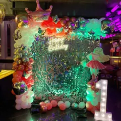 A glamorous mermaid-themed first birthday. This party features a bright sequin wall, a multi-colored balloon arch, a mermaid cutout, and a large, illuminated marquee number '1'.
