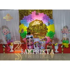 A bright and colorful Cocomelon theme for a second birthday, set against a gold sequin wall. The setup includes character cutouts, a rainbow balloon arch, and a light-up number '2'.