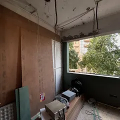 Plywood sheets and construction materials are stacked and ready as a room begins to take shape, with a large window framing the green world outside.