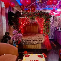 A luxurious and dramatic bedroom setup featuring a four-poster frame completely covered in fresh red roses and greenery. The design is complemented by red balloons, mood lighting, and a candle-lit table.