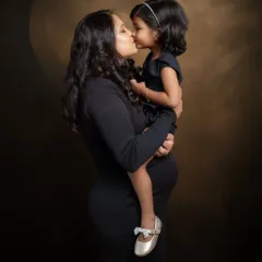 A tender kiss from a soon-to-be big sister. This classic, dark-toned portrait focuses on the intimate connection between mother and child, creating a timeless and emotional piece of art.