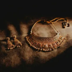 A close-up, artistic shot of the bride's wedding jewelry. The dramatic lighting highlights the intricate design of the gold necklace and earrings, essential elements of the bridal look.