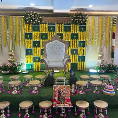 An elaborate and traditional baby shower stage, rich in cultural details. The decor includes a checkered backdrop of green panels and yellow marigolds, a regal white throne, hanging floral garlands, and brass accents, all set on a green turf for an authentic feel.