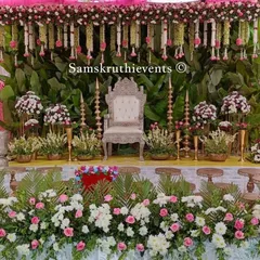 A grand stage setup with a banana leaf backdrop. The decor is enhanced with an ornate silver throne, traditional brass lamps, and a lush border of white and pink flowers.