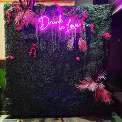 A trendy and edgy photo wall for a cocktail party or sangeet. A 'Drunk in Love' pink neon sign is set against a lush green wall, decorated with tropical pink leaves for a bold and fun statement.