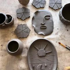 A top-down view of my raw clay creations, including a trinket dish with a sun and moon design, a face plaque, and textured coasters.