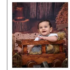 A joyful, open-mouthed smile from a baby boy sitting in a wooden cart. This vintage-themed shoot uses a warm, rustic setting with hay and a barn backdrop to create nostalgic portraits.