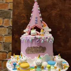 This beautiful unicorn-themed cake features a pastel purple design with a tent-like topper. The cake is surrounded by a group of adorable, happy fondant unicorns sitting on macarons, creating a magical campsite.