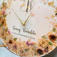 A close-up of a custom 16-inch resin clock. You can see the delicate preserved flowers, gold flakes, and pearls, all set in a milky white resin with the couple's initials.
