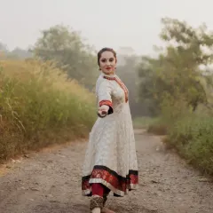 A dynamic shot of the dancer in a powerful Kathak stance. Her direct gaze and outstretched hand invite the viewer into her performance, set against a soft, hazy natural path.