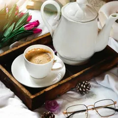 A cozy morning scene with a cup of coffee on a wooden tray in bed. The props, like the open book, glasses, and flowers, help tell a story of a slow, relaxing start to the day.
