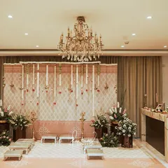 An intimate and traditional engagement setup inside a home. The decor features a backdrop of hanging jasmine garlands and elegant brass lamps, creating a warm and sacred ambiance.