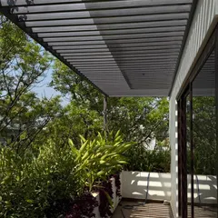 A view from a balcony underneath a metal and glass pergola, where the structure filters the harsh sun and creates a pattern of moving shadows on the floor.
