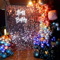 A full view of a dramatic and colorful balloon installation for a birthday party. The organic garland flows from warm oranges and golds to cool silvers, purples, and blacks, creating a striking contrast against the sparkling backdrop.