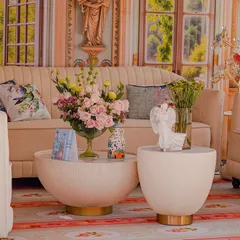 A cozy seating nook from the 'Mint to Be' event, featuring a cream-colored sofa and modern tables. The backdrop is a painted mural of a romantic garden, and the decor includes whimsical angel statues and fresh pink roses.