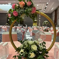 An elegant table centerpiece with a modern design. A golden ring holds a hanging glass orb with a candle, while pink and white roses are arranged at the top and bottom.