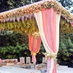 A beautiful pastel-themed outdoor mandap. The soft pink and white drapes, combined with a roof of delicate flowers and greenery, create a light, airy, and romantic setting for a daytime wedding.
