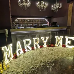 A closer view of the 'MARRY ME' marquee letter decoration. The bright lights stand out beautifully against the dark wood floor and elegant bar setting, creating a high-impact visual.