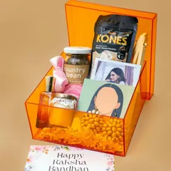 A stylish Rakhi hamper for a sister, presented in a vibrant orange acrylic box. It's filled with coffee, snacks, a scented candle, perfume, and personalized photo items.