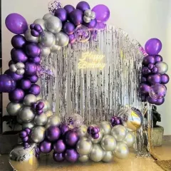 A stylish balloon arch over a silver fringe backdrop. The purple and silver chrome balloons, along with a 'Happy Birthday' neon sign, create a modern and celebratory look.