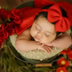 A beautiful two-month-old baby girl sleeping peacefully in a bucket prop. The vibrant red bow and flowers add a lovely pop of color to this newborn portrait.