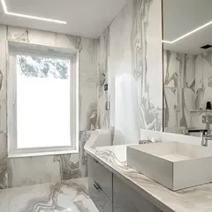 This executive bathroom exemplifies understated luxury with its monochrome palette. The walls and countertop feature Satvario Gold stone with flowing grey veins, creating a cohesive and sophisticated space that is filled with natural light from a large window.