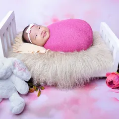 This wide-eyed baby girl is all smiles while lying in a miniature bed, accompanied by a soft toy bunny. The pink floral backdrop adds to the cheerful atmosphere.