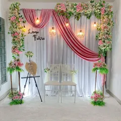 An elegant and romantic engagement stage decoration for an indoor ceremony. This setup includes a white fabric backdrop with gracefully draped pink velvet, framed by lush pink and white floral arrangements and illuminated by warm hanging pendant lights.