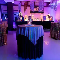 A high-top cocktail table with iridescent linen, reflecting the purple and blue party lights. The centerpiece is a geometric lantern with a candle, adding to the moody and glamorous atmosphere.