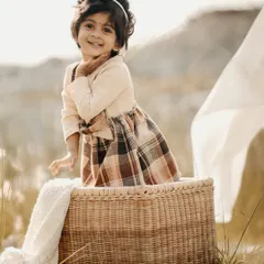 A golden hour portrait of a little girl with a sweet smile, sitting in a wicker basket in a field of tall grass.