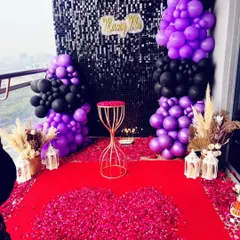 A modern and chic proposal decoration on a balcony. This setup features a black shimmer wall backdrop, bold purple and black balloon arrangements, and a red carpet covered in flower petals for a dramatic effect.