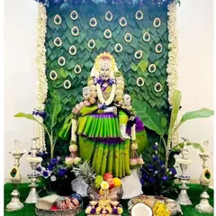 A beautiful decoration for a Varamahalakshmi pooja, with the goddess adorned in a green and purple saree. The backdrop is made of fresh paan leaves arranged in a peacock feather pattern, creating a unique and traditional look.