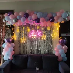 A simple and sweet baby shower decoration designed to fit perfectly in a living room. This setup uses a silver shimmer curtain, a pastel balloon arrangement, and a neon sign placed over the sofa.