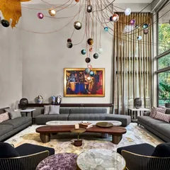 The living room at the Baid Residence is a study in luxurious scale and artistic expression. A custom Bocci light installation cascades from a gold-leafed ceiling, illuminating a space filled with curated furniture and art, including Paresh Maity's work. Large windows frame views of the adjacent forest, seamlessly blending the opulent interior with the natural world outside.
