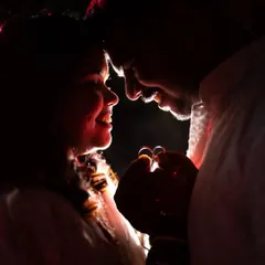 A close-up, dramatically lit photo of the couple holding their engagement rings. The focus is on their hands and the rings, creating a romantic and symbolic image.