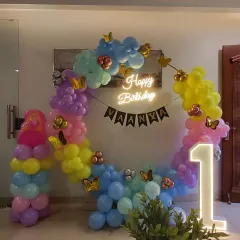 A vibrant butterfly theme for Vaanya's first birthday. This setup uses a circular frame decorated with a rainbow of pastel balloons, a personalized 'Happy Birthday' neon sign, and a large marquee number '1'.