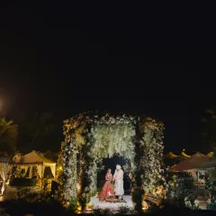 A breathtaking wide shot of a nighttime wedding ceremony. The mandap, covered in white flowers, is set over a pool, its reflection creating a perfect mirror image. The scene is grand, romantic, and utterly magical.