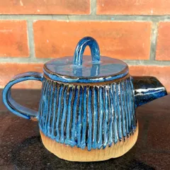 A student's hand-built teapot with a carved vertical line texture and a rich blue glaze. The unglazed base provides a beautiful contrast.