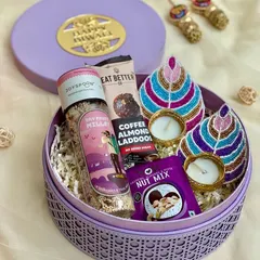A round rattan box in a lovely lilac color, filled with healthy treats and hand-painted diyas for a unique Diwali gift.