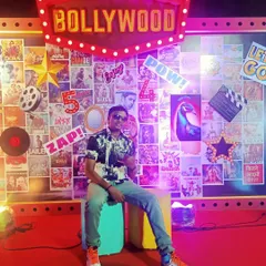 Posing in front of a Bollywood-themed backdrop. I am your go-to DJ for desi parties, with an extensive library of Bollywood and regional hits.
