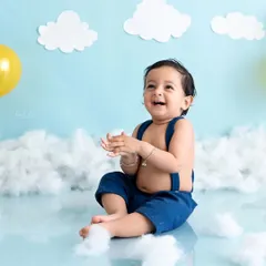 A happy baby sits amidst a sky-themed backdrop with fluffy clouds and balloons, clapping his hands with joy.