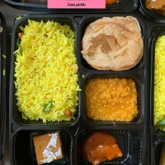 A close-up of a 'No Greens, No Veggies' meal box. This thali includes Puri, Mogar Dal, Lemon Rice, Besan Chikki, and lime pickle, ensuring a complete and tasty meal.