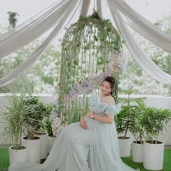 Seated gracefully, this mom-to-be shows how comfortable and beautiful our rental gowns are. The garden-themed studio setup with the birdcage adds a whimsical touch to the maternity photo.