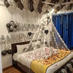 An elegant anniversary surprise using a black and silver color scheme. The setup includes a canopy with fairy lights, "Happy Anniversary" foil balloons, and a swan towel arrangement on a bed of rose petals.