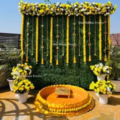 A classic Haldi setup for the groom on a terrace, featuring a green wall backdrop with hanging marigold strings and bell tassels. The look is completed with potted marigolds and a traditional 'urli' for the ceremony.
