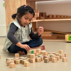 Sitting on the floor, a young girl carefully stacks small wooden cubes. This activity requires precision and patience, helping to refine her fine motor skills and concentration.