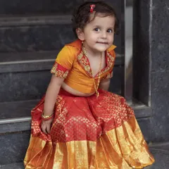 Our cute little client seated on the stairs, looking adorable in her custom orange and red brocade Langa Dhawani.