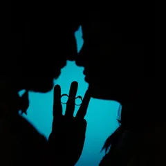 A creative silhouette of the couple, their hands forming a 'V' to frame their wedding rings against a bright blue background. A unique and artistic engagement shot.