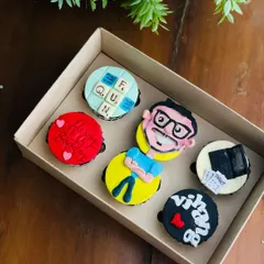 A box of personalized cupcakes with custom toppers, including a hand-painted portrait, Scrabble tiles, and other hobby-related designs.