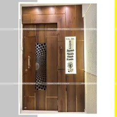 A modern entrance door design for a 1BHK apartment. The door features a unique metal jali insert and is surrounded by wooden paneling with brass inlays, creating a sophisticated and contemporary look.
